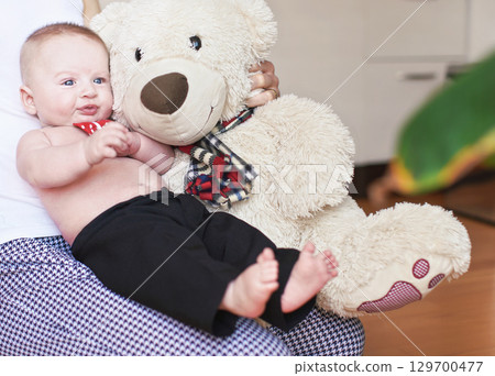 Four months old baby boy sitting on her mother knees, large plush teddy bear next to him 129700477