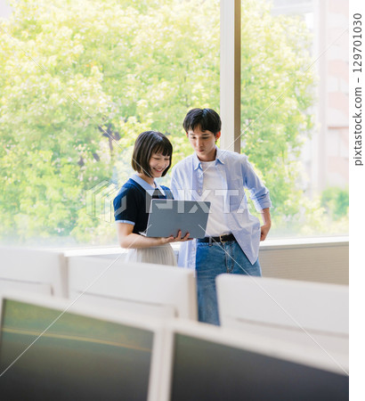 Male and female university students having a conversation by the window with a laptop Male and female university students having a conversation by the window with a laptop 129701030