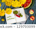 Fall still life with an open book labeled Hello, Autumn surrounded by bright yellow flowers, apples, juicy watermelon, and red berries on wooden table 129703099