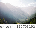 Summer view into a valley in the Austrian Alps. Sunlight shines over the landscape, with forested mountains on the sides and a winding road and river at the bottom of the valley. 129703192