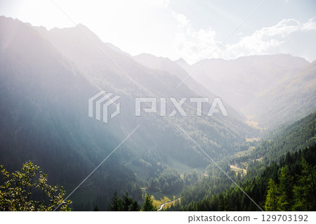 Summer view into a valley in the Austrian Alps. Sunlight shines over the landscape, with forested mountains on the sides and a winding road and river at the bottom of the valley. 129703192