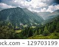 Summer view into a valley in the Austrian Alps. Sunlight shines over the landscape, with forested mountains on the sides and a winding road and river at the bottom of the valley. 129703199