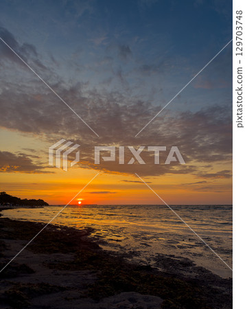 Sunset over the tranquil ocean with colorful clouds illuminating the horizon near a sandy beach during evening hours 129703748