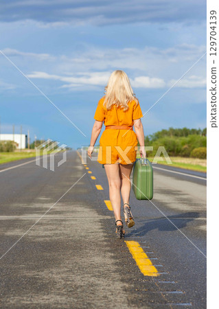 Blonde woman with a green retro suitcase walking on the highway. Blonde woman with a green retro suitcase walking on the highway. 129704139