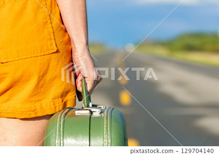 Woman's hand is holding a retro green suitcase and walking on a highway. Woman's hand is holding a retro green suitcase and walking on a highway. 129704140