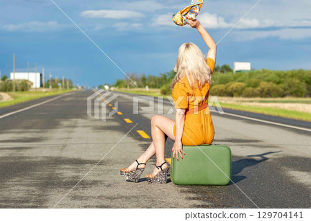 Blonde woman is sitting on a retro suitcase on a rural highway. Blonde woman is sitting on a retro suitcase on a rural highway. 129704141