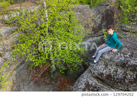 Boy is sitting on big Rocks formation in the green forest. 129704202
