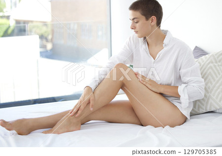 Woman, legs and moisturizer for healthy skincare, cosmetics or body care relaxing on bed at home. Female rubbing her leg for silky smooth, soft or nourished skin treatment sitting in the bedroom 129705385