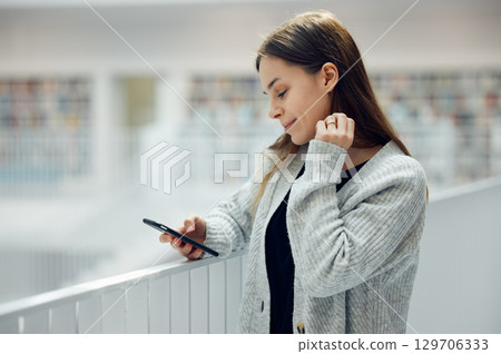 Woman, phone and smile for social media, networking or chatting in communication at a library. Happy female smiling for texting, research or browsing for books on smartphone at big book store or mall Woman, phone and smile for social media, networking or chatting in communication at a library. Happy female smiling for texting, research or browsing for books on smartphone at big book store or mall 129706333