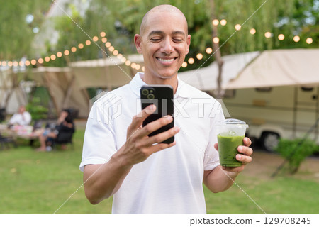 Bald man at campsite with camper van having healthy green detox juice drink and using phone Bald man at campsite with camper van having healthy green detox juice drink and using phone 129708245