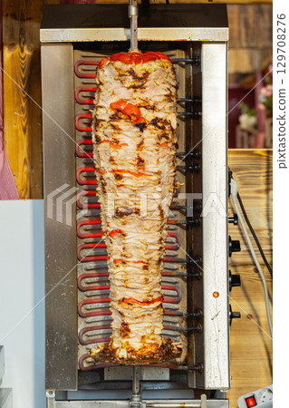 Vertical rotisserie with seasoned meat and vegetables cooking in a commercial kitchen setting Vertical rotisserie with seasoned meat and vegetables cooking in a commercial kitchen setting 129708276