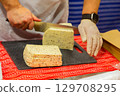 Hands slicing cheese on a black cutting board with colorful textile underneath for culinary presentation 129708295