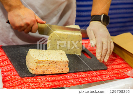 Hands slicing cheese on a black cutting board with colorful textile underneath for culinary presentation 129708295