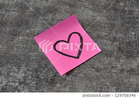 A pink sticky note with a heart sketch rests on a gray surface A pink sticky note with a heart sketch rests on a gray surface 129708346