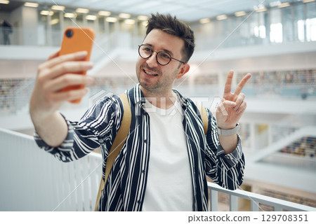 College student, man and library selfie at campus with phone, peace hand and smile on social media. Gen z, university student and smartphone app for social network, happiness and studying for success College student, man and library selfie at campus with phone, peace hand and smile on social media. Gen z, university student and smartphone app for social network, happiness and studying for success 129708351