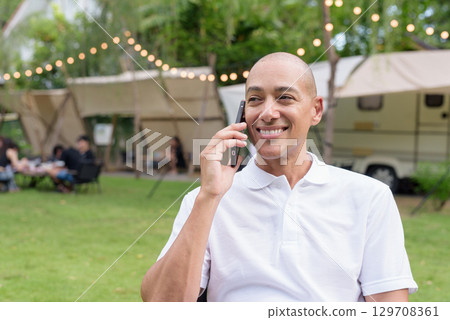Confident bald man relaxing at campsite with camper van using phone 129708361