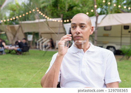 Confident bald man relaxing at campsite with camper van using phone Confident bald man relaxing at campsite with camper van using phone 129708365