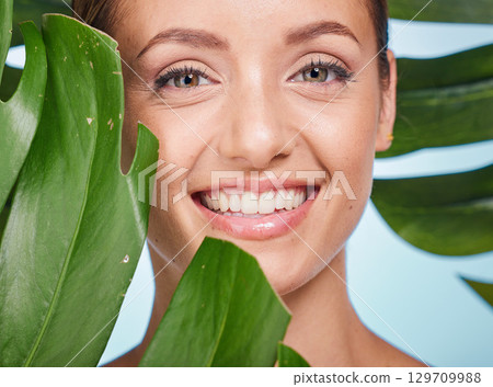 Portrait, skincare and palm leaf with a model woman posing in studio on blue background for beauty. Face, skin or nature with an attractive young female standing behind a plant for natural treatment Portrait, skincare and palm leaf with a model woman posing in studio on blue background for beauty. Face, skin or nature with an attractive young female standing behind a plant for natural treatment 129709988