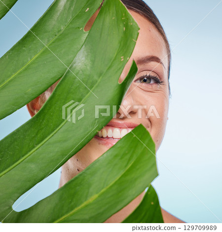 Portrait, skincare and palm leaf with a model woman posing in studio on blue background for beauty. Face, skin or nature with an attractive young female standing behind a plant for natural treatment Portrait, skincare and palm leaf with a model woman posing in studio on blue background for beauty. Face, skin or nature with an attractive young female standing behind a plant for natural treatment 129709989