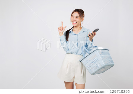 Smiling Asian woman pointing finger to empty space and holding mobile phone with shopping basket excited about online shopping, grocery market, and digital commerce. isolated studio white background 129710295
