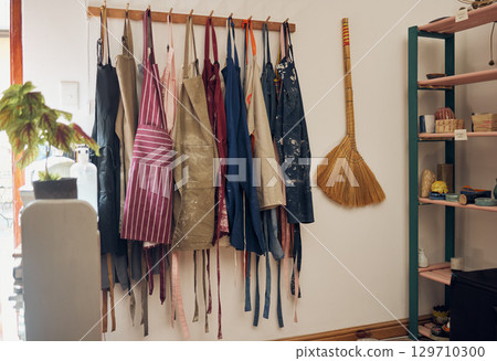 Apron, hanging and art shop with a broom in an empty workshop or studio against still life wall for design. Creative, pottery and clothing with a group of aprons on a rack in a small business startup Apron, hanging and art shop with a broom in an empty workshop or studio against still life wall for design. Creative, pottery and clothing with a group of aprons on a rack in a small business startup 129710300