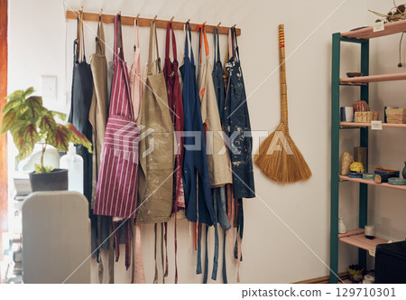 Clothes rack, apron and paint with a broom in an empty workshop or studio against still life wall for design. Creative, art and clothing with a group of aprons hanging in a small business startup 129710301