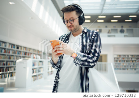 Phone music, headphones and student in library, university, or college research on mental health, wellness and education. Bookshelf, knowledge and creative, geek man listening to audio on mobile app 129710338