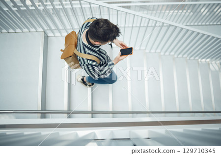 Phone, stairs and education with a student man walking down a staircase at university or college. Mobile, study and top view with a male pupil taking steps to get to class for learning or development 129710345