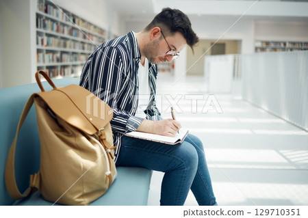 Library, student and college man writing ideas for educational task in notebook with focus. Learning, knowledge and education of smart university guy thinking of assignment with concentration. 129710351