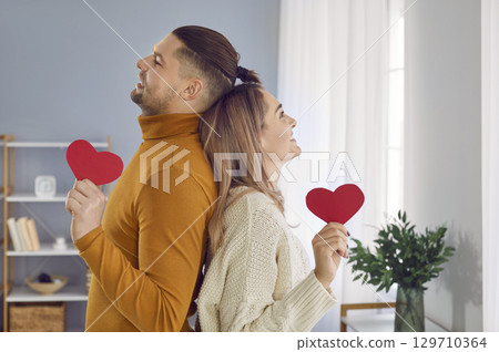 Portrait of young man and woman couple standing back to back and holding red hearts in hands. 129710364