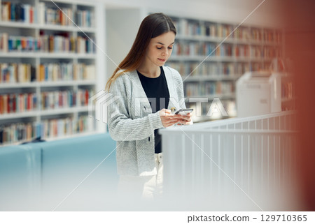 Library, phone and woman student networking on social media, mobile app or internet in college. Bookstore, knowledge and young female typing or reading a text message on her cellphone at university. 129710365