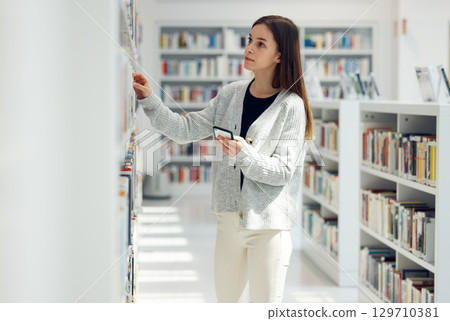 Library, book choice and student with phone in university, college or school. Learning, education scholarship or woman holding mobile looking for books by bookshelf for reading, knowledge or studying Library, book choice and student with phone in university, college or school. Learning, education scholarship or woman holding mobile looking for books by bookshelf for reading, knowledge or studying 129710381
