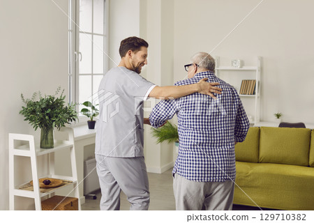 Young nurse man helping a senior grey-haired patient to walk at home in rehab. 129710382