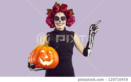 Beautiful young woman with face painted for Halloween holding scary pumpkin 129710459