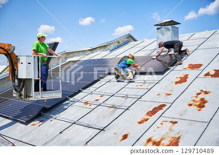 Workers building solar panel system on metal rooftop of house with assistance of crane lift. Three men installers installing photovoltaic solar module outdoors. Renewable energy generation concept. 129710489
