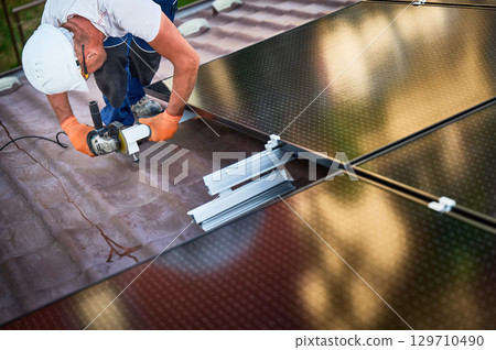 Worker installing or maintaining photovoltaic solar panel system on roof of house. Technician in helmet using grinder on metal part of the panel's mounting structure. Concept renewable energy. 129710490