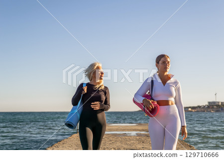 Women Yoga Mat Beach Walk - Two women walk along a pier with yoga mats after an outdoor yoga session. 129710666