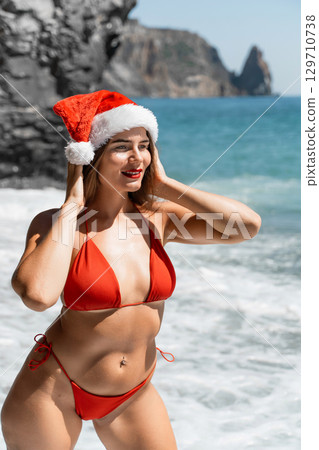 A woman in Santa hat on the seashore, dressed in a red swimsuit. New Year's celebration in a hot country A woman in Santa hat on the seashore, dressed in a red swimsuit. New Year's celebration in a hot country 129710738