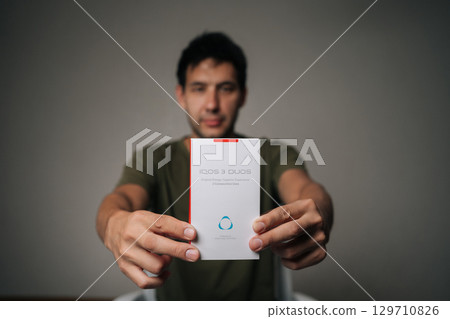 Ufa, Russia - 21 July 2025. Blurred portrait of influencer male holding and presenting new IQOC 3 duos tobacco heating system, innovative electronic cigarette for modern smokers seeking harm reduction Ufa, Russia - 21 July 2025. Blurred portrait of influencer male holding and presenting new IQOC 3 duos tobacco heating system, innovative electronic cigarette for modern smokers seeking harm reduction 129710826