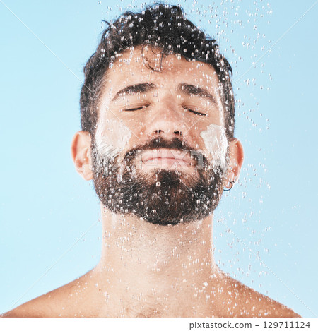 Water, man with face and cream for cleaning in shower with hygiene, grooming and skincare against blue studio background. Clean, model with water drops and facial, natural treatment and cosmetic care 129711124