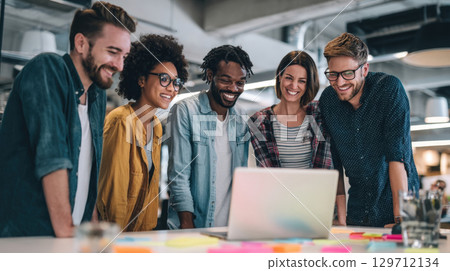 A group of smiling, diverse young professionals enthusiastically collaborate and brainstorm, gathered around a laptop in a bright, contemporary office environment 129712134