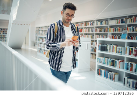 Education, social media and man with a phone in a library for communication, wifi connection and online chat at college. University, learning and student with a mobile for research and typing email 129712386