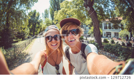 A cheerful young couple, wearing sunglasses and casual attire, smiles brightly for a selfie while enjoying a sunny day outdoors in park like setting. Ideal for themes of summer, travel, and friendship 129712726
