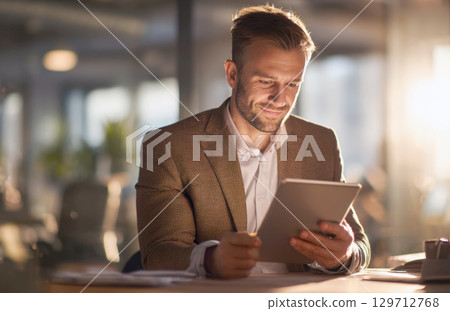 A well-groomed businessman in a stylish suit jacket attentively interacts with a digital tablet in a modern office setting, illuminated by warm, ambient light. Perfect for business, technology, 129712768