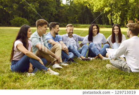 Man taking interview from a group of a young people sitting in the summer park. 129713019