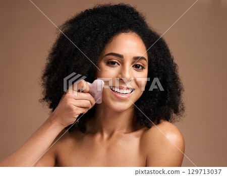Woman, studio portrait and crystal guasha for skincare, smile or cosmetic facial massage by backdrop. Black woman, model and self care with rose quartz stone for face glow, skin wellness or aesthetic 129713037