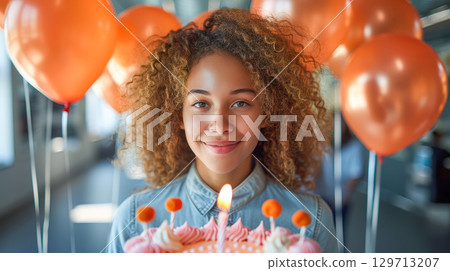 Young girl with curly hair holding a birthday cake surrounded by festive balloons and celebration atmosphere 129713207