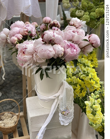 Bouquet of pink peonies in a white vase surrounded by yellow flowers at an outdoor market 129713284