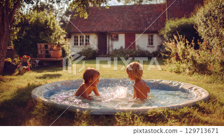 Two happy children are seen from behind, playing, splashing in small inflatable swimming pool in backyard. Warm light of late afternoon illuminates the scene, with house, lush greenery in background 129713671