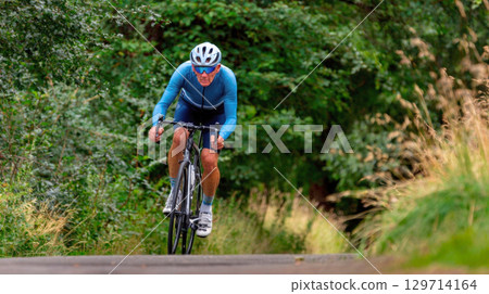A focused man in athletic cycling gear riding a road bike up a paved hill, surrounded by lush green foliage. Perfect for themes of sport, fitness, determination, and outdoor activity A focused man in athletic cycling gear riding a road bike up a paved hill, surrounded by lush green foliage. Perfect for themes of sport, fitness, determination, and outdoor activity 129714164